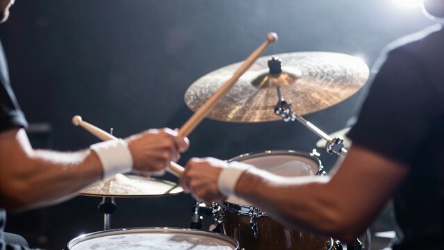 A drummer plays drums with passion under dramatic stage lighting, hands gripping sticks mid-motion near cymbals and snare drum.