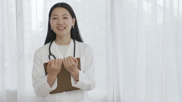 Young Japanese female doctor holding binder smiling by window