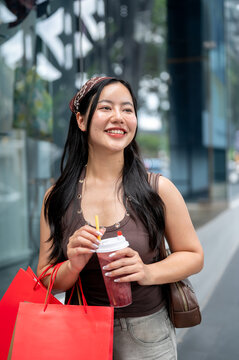 Young Asian woman holding berry smoothie and shopping bags in outdoor retail district, happy lifestyle portrait