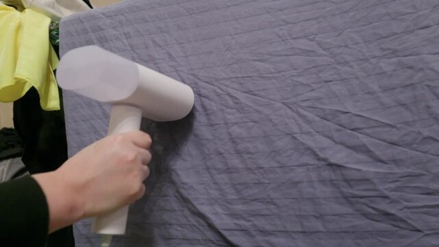 Person in yellow safety vest operating portable garment steamer on gray fabric, demonstrating professional cleaning technique with visible steam application.