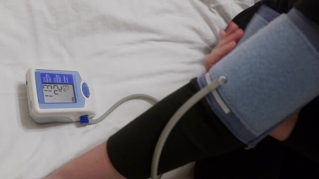 Individual checking blood pressure using portable digital monitor with cuff around arm, displaying measurement on blue screen display against white background.