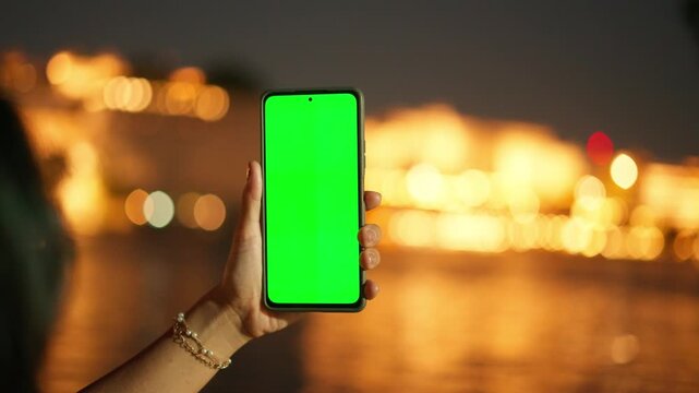 4K shot of woman hand holding mobile phone with green screen and tap on screen, blurred lighting of Udaipur city in background. Green screen mockup template of Mobile phone, Chroma key.