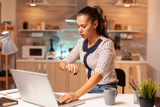 Late deadline keeps a businesswoman working beyond normal hours as she checks the time beside her laptop, checking the time highlights pressure as the deadline approaches.