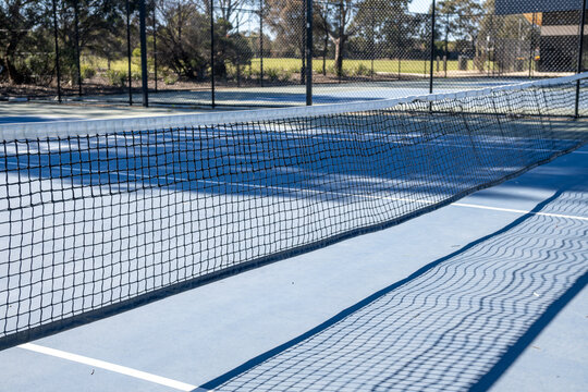 Mesh tennis net across a blue hardcourt surface, with clear net shadows cast on the ground surrounded by chain-link fencing&nbsp;in a suburban Australian outdoor court.