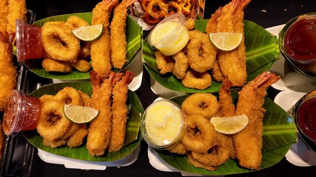 Assorted deep fried seafood platters served with crispy breaded shrimp, squid rings, dipping sauces and lemon wedges at a Thai street food market. Delicious seafood takeaway meals presented on banana 