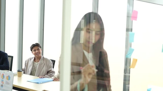 Businesswoman writing ideas on glass board during team meeting, project planning, brainstorming, office collaboration, business strategy, corporate teamwork, innovation workshop.