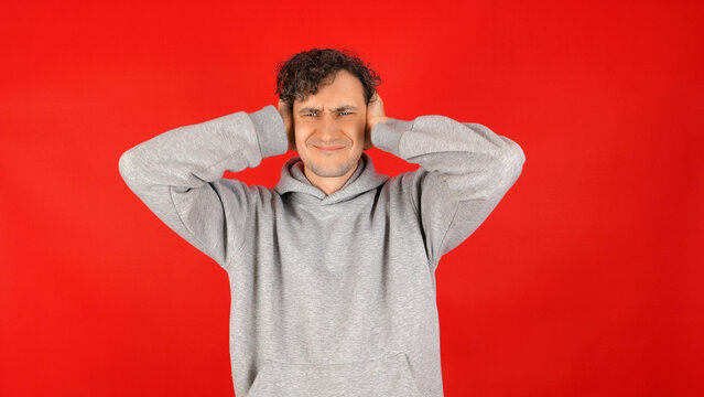 Stressed young man in grey hoodie holding head with both hands, grimacing in frustration against solid red studio background, overwhelmed by trouble and pressure.