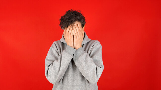 Young curly-haired man in grey hoodie covering his face with both hands against vibrant red studio background expressing embarrassment frustration failure and emotional distress.