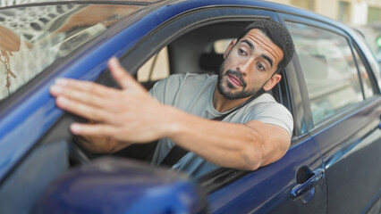 Naklejka premium Man in blue sedan leaning out of car with bare forearm reaching and gesturing through open window on street; anger confrontation.