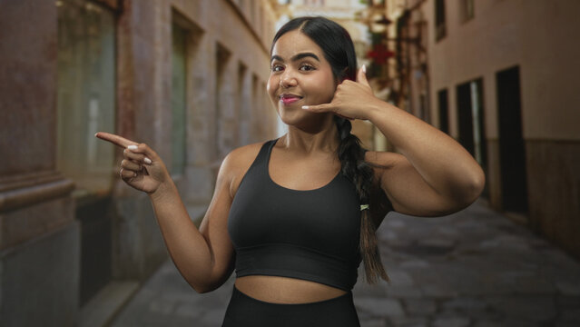 Young sporty hispanic woman points finger and makes phone hand gesture wearing black sports bra on narrow cobblestone street; confidence invitation.