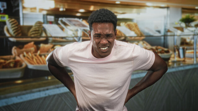 Man in pink tshirt clutching stomach with hands amid bakery pastries by bread display at building counter; pain nausea urgency.