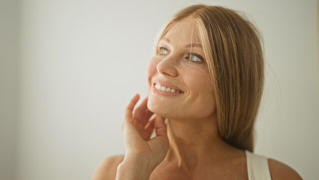 Woman young blonde hand to cheek smiling toward soft natural light against white wall in studio; quiet serenity.