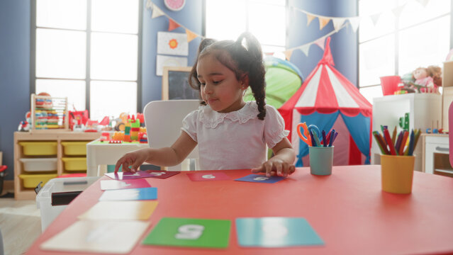 Girl placing number cards with hand on a red activity table, sitting in a bright playroom with tent and toy storage in building; concentration learning.