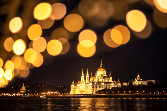 Illuminated Hungarian parliament building stands majestically along the dark riverbank at night featuring warm glowing golden bokeh highlights created by out of focus city street lights