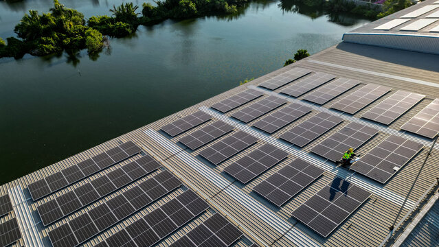 Aerial view of a lone solar technician on a rooftop array with a canal and tropical trees in the warm evening, showing clean renewable energy generation at an industrial site in Thailand.