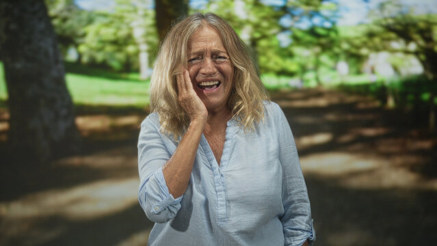 Senior woman holding cheek with hand in forest park, eyes closed and wincing from toothache while wearing light blue blouse and standing on shaded path; pain vulnerability.