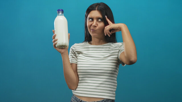 Woman holding milk bottle and pointing finger to temple in studio wearing striped shirt with a wince expression; playful skepticism.