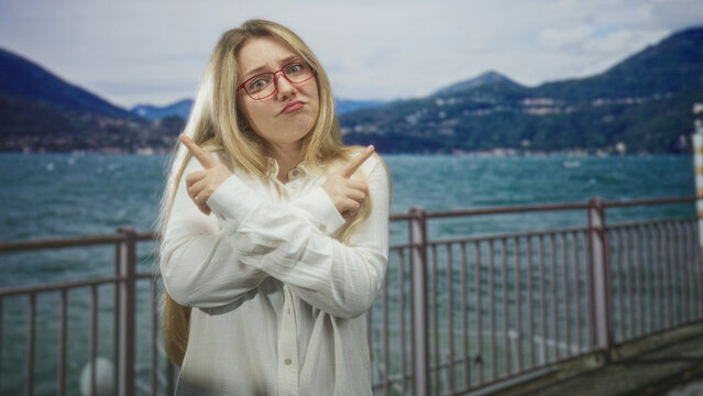 Woman, blonde, shrugging with hands at a lakeside pier in a white shirt and red glasses, looking puzzled and slightly resigned; uncertainty pondering.