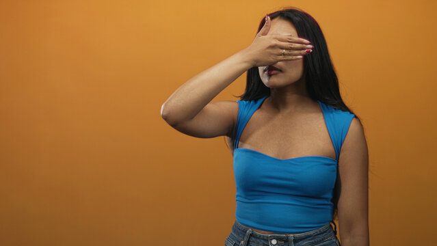 Woman covering eyes with hand, palm over face, wearing blue strap top and jeans, long brunette hair visible against orange studio wall; shyness.