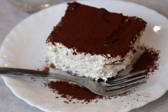 Porzione di tiramis&ugrave; artigianale servita su un piatto bianco con polvere di cacao e un cucchiaino da dessert sul tavolo di un ristorante