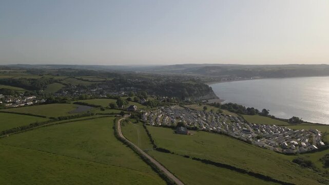 Aerial drone panorama of Seaton Holiday Caravan Park with views across the surrounding landscape of Devon. Drone moving forward over the green fields in direction of the caravan park.
