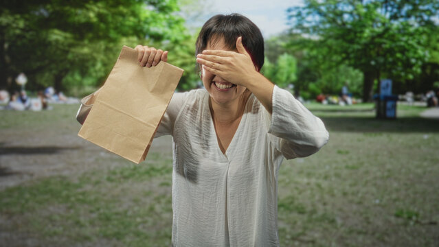 Woman holding a paper bag with hand covering eyes and smiling amid trees in a forest picnic setting; joy surprise.