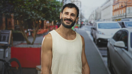 Fototapeta premium Man with bare arms smiling and tilting head on a busy street wearing a sleeveless knit tanktop; contentment easygoing summer.