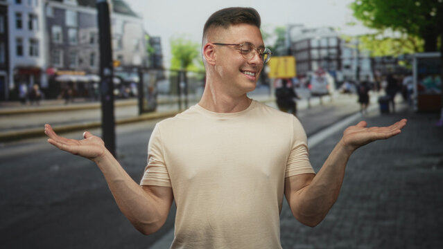 Man shrugging with palms up on a city street wearing glasses and beige tshirt; nonchalance indecision.