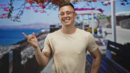 Fototapeta premium Young man smiling makes rock hand gesture with bare hand and forearm in a studio seaside set with pergola backdrop; playful ease.