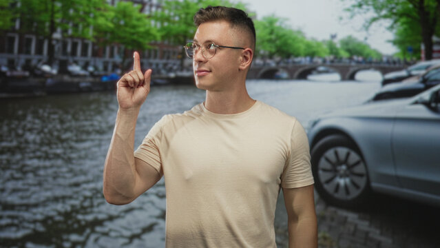 Man wearing glasses shows thumbs down with right hand beside canal and parked car on street; disapproval critique.