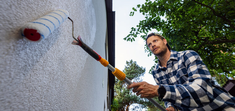 House painter applying protective coating on house exterior wall with roller. Facade protection against algae, moss and fungal