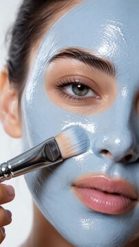 Beautiful young woman applying blue clay facial mask with a cosmetic brush. Close-up view of skincare routine, facial treatment, and spa procedure for clean skin and wellness