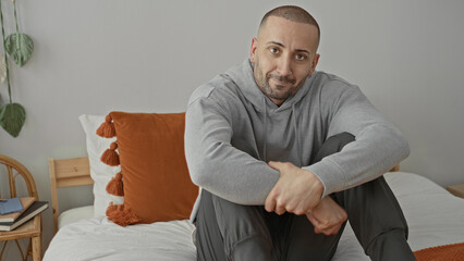 Fototapeta premium Man sitting on bed in gray hoodie, hands clasped over knee, slight smile, studio; calm quiet reflection.