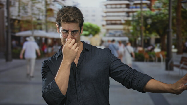 Man pinches nose with hand and holds out other arm while wearing glasses and dark shirt on busy street plaza with blurred pedestrians; discomfort.