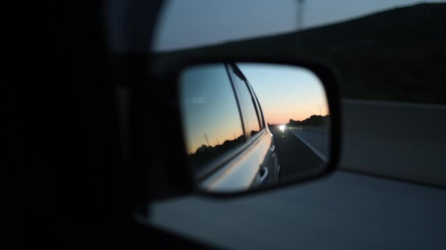 Car side mirror reflects an open highway at night as travelers continue a summer road trip with headlights approaching behind and soft sunset colors fading across the sky