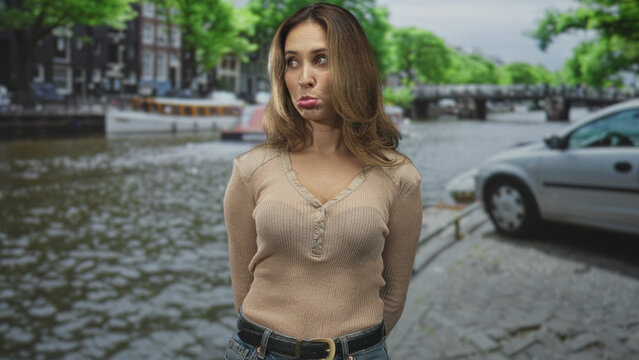 Woman pouting lips with visible cleavage standing beside a canal street and parked car, hands behind back; melancholy reflection.