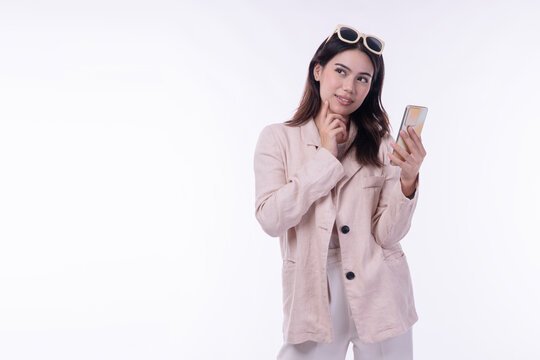 Thoughtful young Asian businesswoman in beige blazer with sunglasses on head holding smartphone and looking away on white background with copy space. Female pondering or thinking concept.