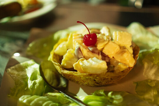 Close up of delicious fruit salad in crispy edible taro basket topped with red cherry and creamy dressing on plate with lettuce. Healthy vegetarian appetizer or dessert for catering event.
