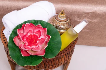 Thai massage and spa setting with pink lotus flower, white towel, traditional ceramic jar and yellow essential oil bottle in woven basket. Relaxing aromatherapy and natural wellness treatment concept. © kaew6566