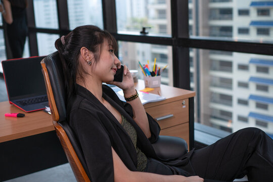 Relaxed young Asian businesswoman in black suit talking on smartphone and looking out window at city view while sitting in office chair. Corporate executive taking a break and communication concept.