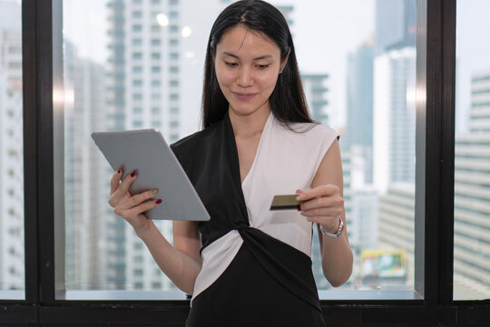 Beautiful young Asian businesswoman holding digital tablet and credit card for online shopping standing by the window with modern city view. Digital payment and corporate lifestyle concept.