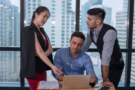 Diverse business team of three people discussing project strategy on laptop in modern office with city skyline view. Corporate colleagues collaborating and analyzing data concept.