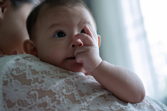 Close up portrait of cute little Asian baby infant resting on mother shoulder looking away near window. Tender moments of motherhood, childcare and family bonding concept.