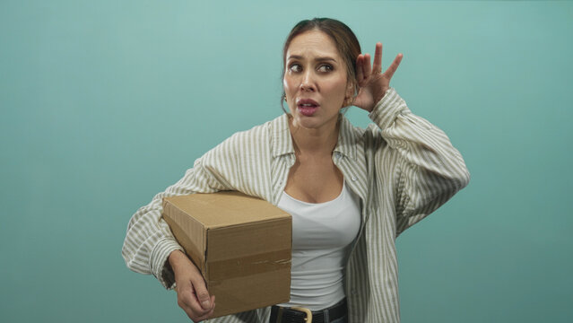 Woman holding cardboard box, hand cupping ear while peering aside, wearing striped shirt and white tank top in studio teal backdrop; concern listening.