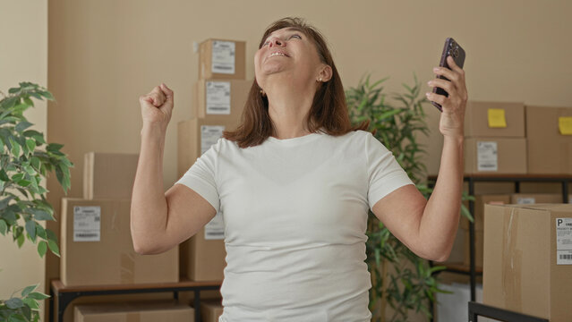 Woman raising fist and holding smartphone beside stacked cardboard boxes in a building; small business triumph.