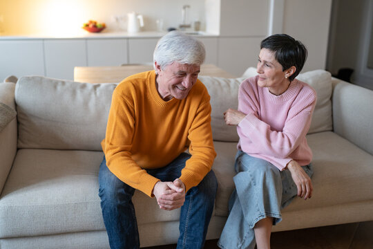 Relaxed elderly couple husband and wife sitting on couch at home joking and having fun together, cheerful senior partners in joyful conversation, family relationships and bonding, emotional connection
