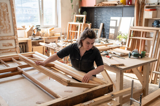 Confident woman in carpentry workshop works at workbench with vintage wooden window frame, female crafter carefully adjusts and checks parts of antique wooden window frames before reconstruction.