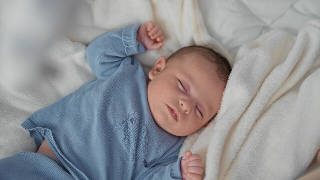 Boy sleeping with face visible on a bed in a nursery, wrapped in blue knit sweater and plush white blanket with arms relaxed; calm gentle rest.