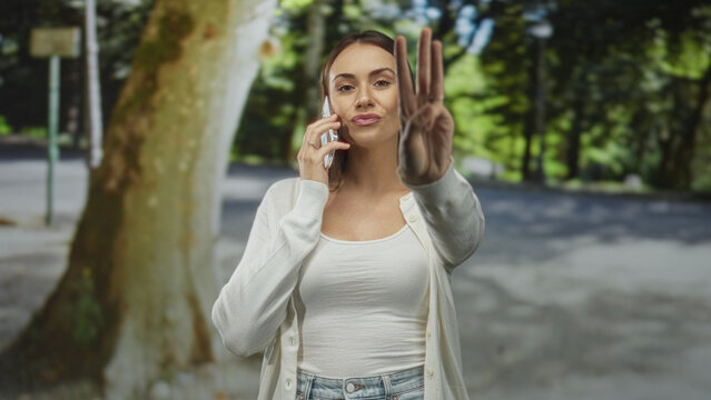 Woman on a mobile phone points finger toward camera on a street beside a bus stop and park trees; assertive conversation.