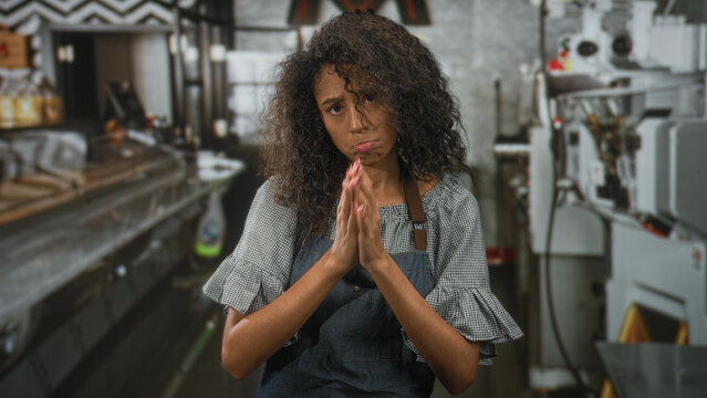 Woman waitress with curly hair and apron, hands clasped pleading in a busy restaurant kitchen, pout expression and closed eyes; apology fatigue.